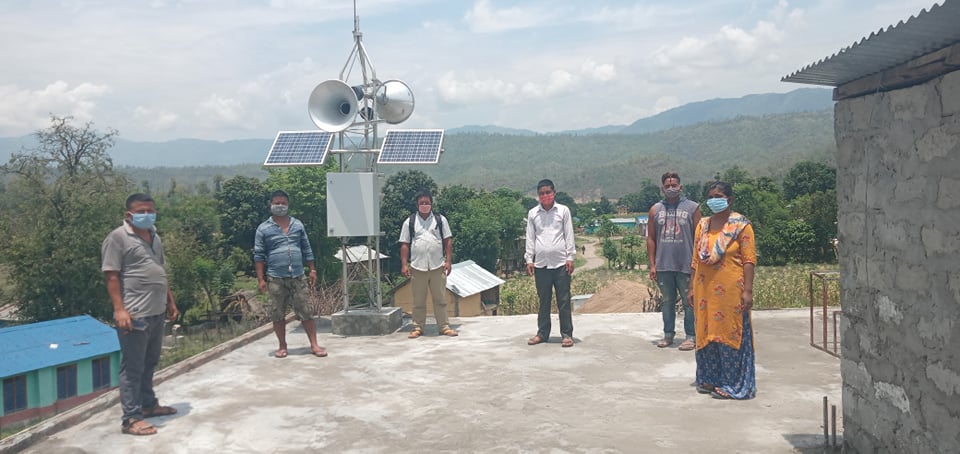 Community members, wearing face masks, standing by the early warning system as the system is tested