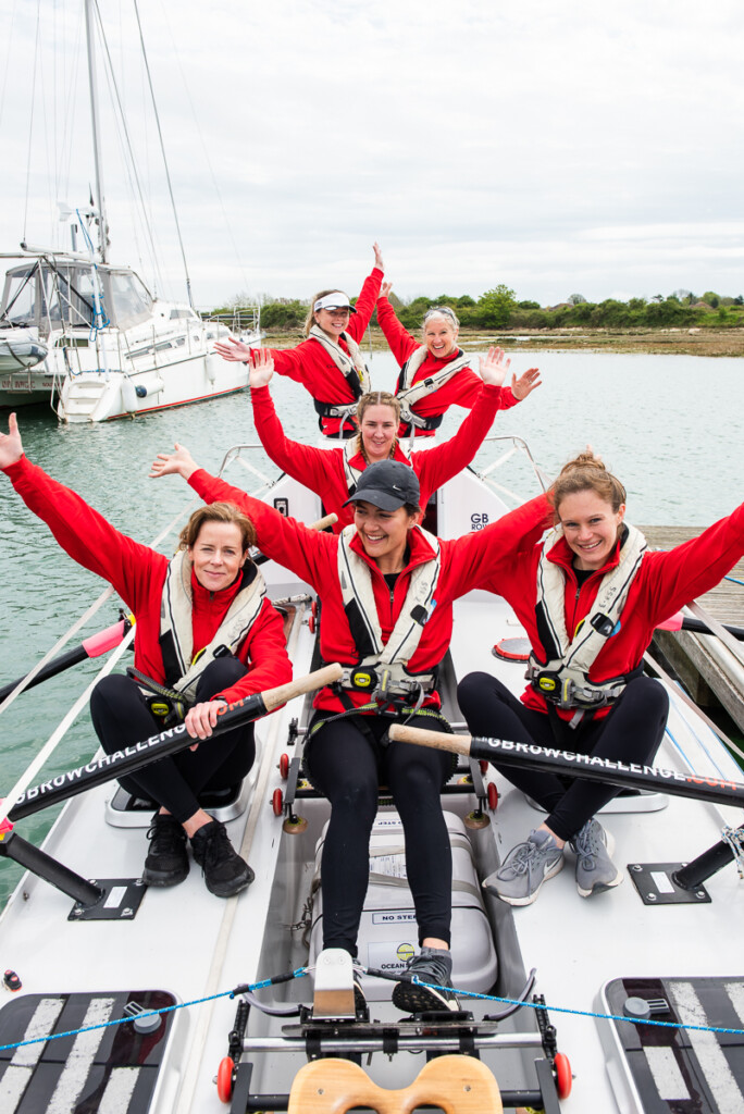 The All Systems Row team: all six ladies are sitting in their rowing boat, on the water, smiling and with their arms raised in celebration.