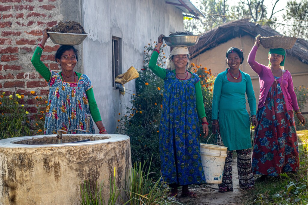 Four smiling Nepali women, three of them are carrying items on their head, one a bucket in her hand. All are benefitting from a Renewable World biogas project.