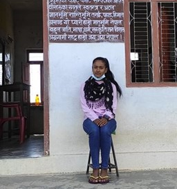 A female Nepali school student is sitting on a stool outside her classroom