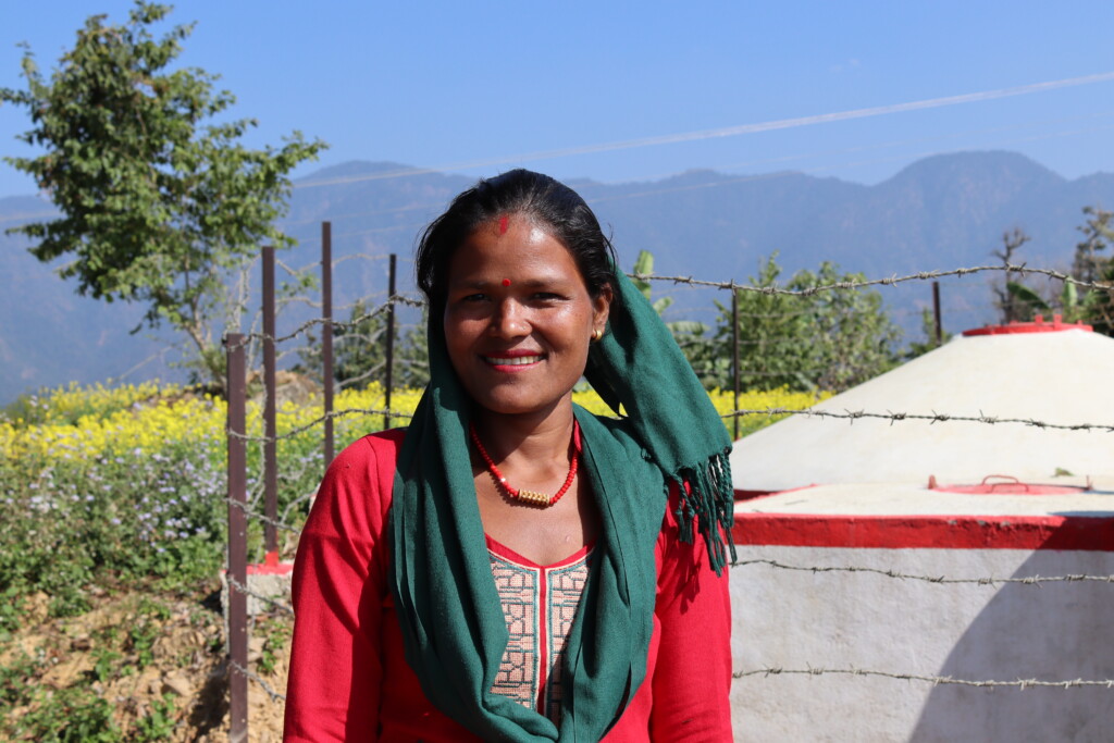 A Nepali woman smiling at the camera, she is standing next to a water storage tank, in the distance you can see tall mountains